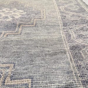 May include: Close-up of a blue and gray patterned rug. The rug features a geometric design with a border. The color palette includes shades of blue, gray, and beige. The rug has a textured surface.