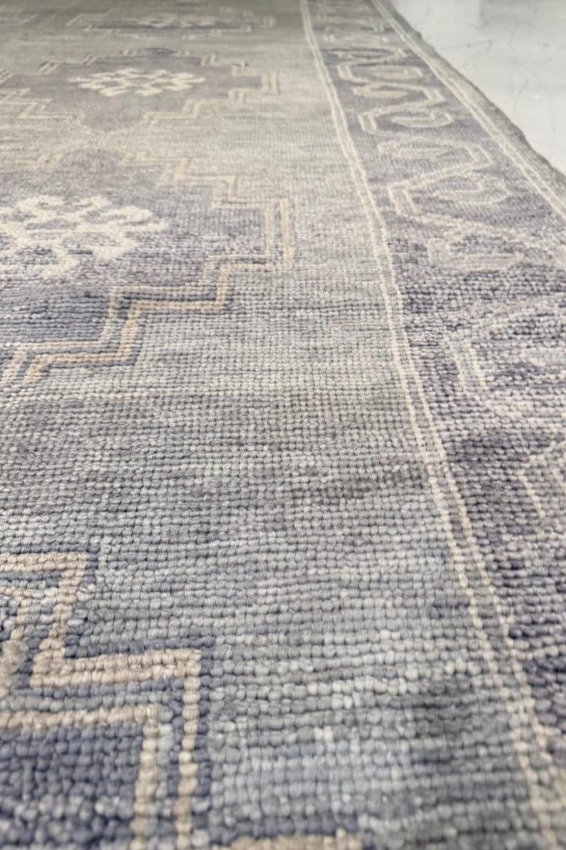 May include: Close-up of a blue and gray patterned rug. The rug features a geometric design with a border. The color palette includes shades of blue, gray, and beige. The rug has a textured surface.