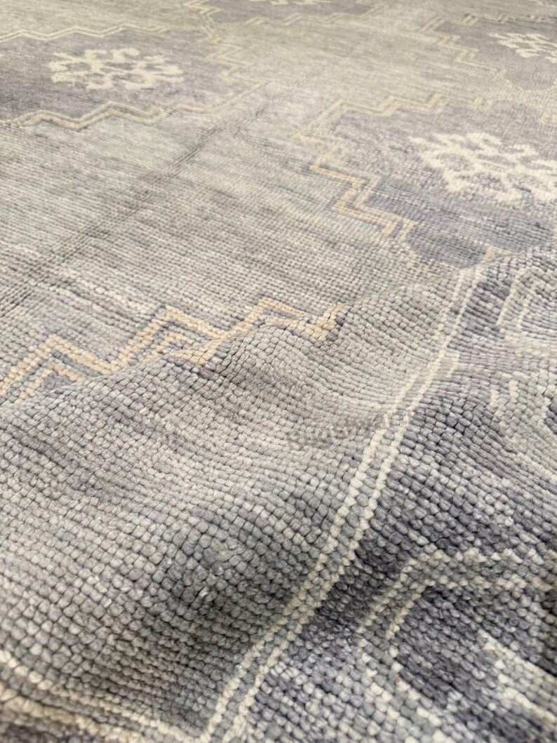 May include: Close-up of a textured, light blue rug with geometric and floral patterns in off-white and beige. The rug's surface has a soft, plush appearance, with visible woven details. The design features a repeating pattern.