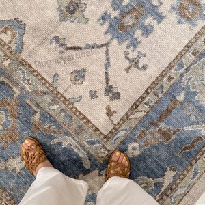 May include: A beige and blue patterned rug with a geometric design. The rug features a central field with floral motifs and a border with a repeating pattern. The image also shows a person wearing brown sandals and white pants.