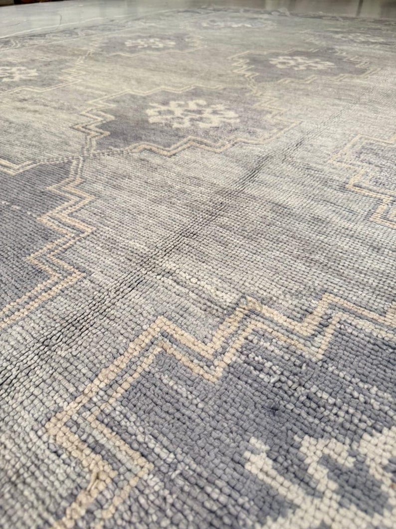 May include: A close-up view of a large, patterned rug. The rug features a distressed design with a combination of light gray, dark gray, and beige colors. The pattern includes geometric shapes and a zig-zag border.