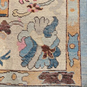 May include: Close-up of a beige rug with a floral pattern in pink, blue, and brown. The rug has a light blue border and a fringe along the bottom edge. The design features stylized flowers and geometric shapes.