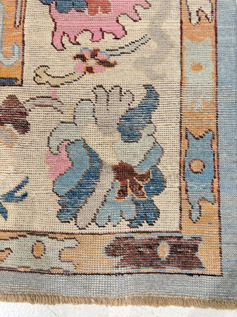 May include: Close-up of a beige rug with a floral pattern in pink, blue, and brown. The rug has a light blue border and a fringe along the bottom edge. The design features stylized flowers and geometric shapes.
