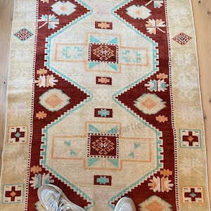 May include: A rectangular rug with a central cream-colored field and a geometric pattern in shades of red, teal, and orange. The rug's border is a deep red, and the design includes diamond and floral motifs. The rug is placed on a light-colored wooden floor.