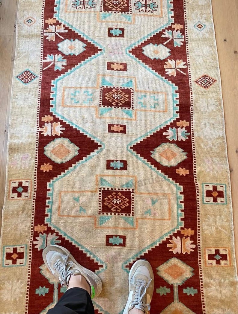 May include: A rectangular rug with a central cream-colored field and a geometric pattern in shades of red, teal, and orange. The rug's border is a deep red, and the design includes diamond and floral motifs. The rug is placed on a light-colored wooden floor.