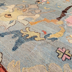 May include: Close-up of a vintage rug with a light blue background and intricate patterns. The rug features designs in shades of yellow, pink, brown, and blue. The texture of the rug is visible, showcasing the woven construction and detailed artistry.
