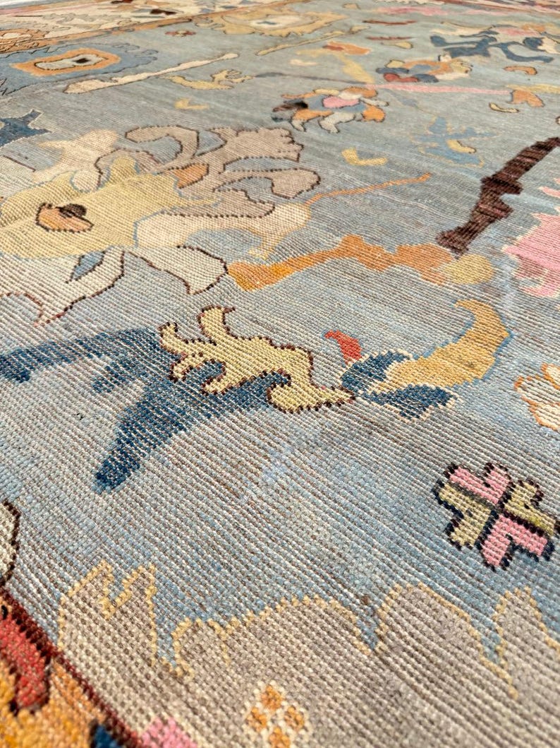 May include: Close-up of a vintage rug with a light blue background and intricate patterns. The rug features designs in shades of yellow, pink, brown, and blue. The texture of the rug is visible, showcasing the woven construction and detailed artistry.