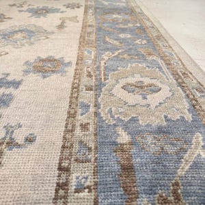 May include: A rectangular rug with a cream-colored field and a blue border. The rug features floral and geometric patterns in shades of blue, brown, and beige. The rug is made of a woven material and appears to be a traditional style.