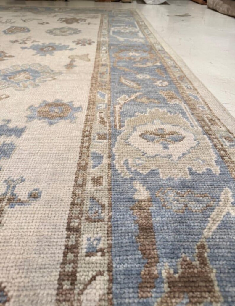 May include: A rectangular rug with a cream-colored field and a blue border. The rug features floral and geometric patterns in shades of blue, brown, and beige. The rug is made of a woven material and appears to be a traditional style.