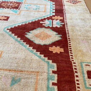 May include: Close-up of a beige and red patterned rug. The rug features geometric designs in shades of red, turquoise, and peach. The rug's border is a deep red, and the overall texture appears woven.