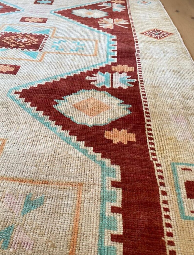 May include: Close-up of a beige and red patterned rug. The rug features geometric designs in shades of red, turquoise, and peach. The rug's border is a deep red, and the overall texture appears woven.