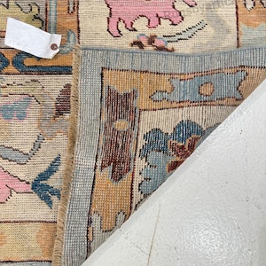 May include: A section of a vintage rug with a detailed floral pattern. The rug features a cream-colored background with blue, pink, and gold floral motifs. The border is a light blue with a geometric design. A tag is attached to the rug.
