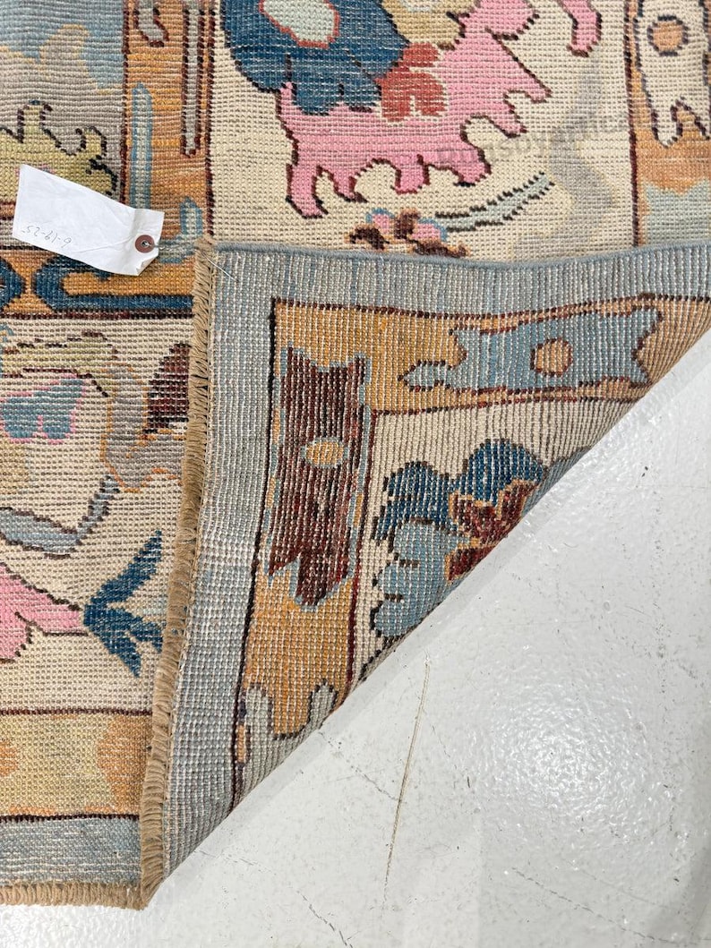 May include: A section of a vintage rug with a detailed floral pattern. The rug features a cream-colored background with blue, pink, and gold floral motifs. The border is a light blue with a geometric design. A tag is attached to the rug.