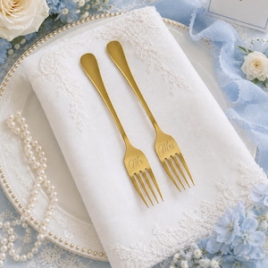 May include: Two gold-toned metal forks resting on a white fabric napkin with lace trim. One fork is engraved with the text Mr and the other with the text Mrs in a cursive script. The forks have long, slender handles and four tines each.