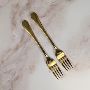 May include: Two gold-colored forks with the words "Mrs" and "Mr" engraved on the tines. The forks have long, slender handles and are set against a marble-patterned surface.