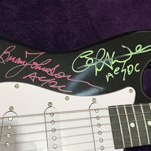 Pu&ograve; includere: Chitarra elettrica nera con battipenna bianco e hardware argentato. La chitarra presenta firme con inchiostro rosa e verde, tra cui "Brian Johnson AC/DC" e "AC/DC". Il corpo della chitarra &egrave; nero e il battipenna &egrave; bianco.
