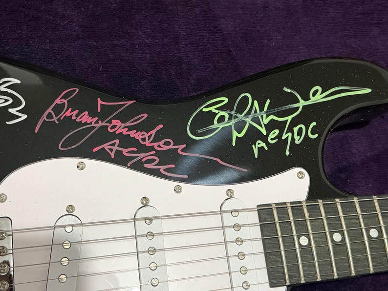 Pu&ograve; includere: Chitarra elettrica nera con battipenna bianco e hardware argentato. La chitarra presenta firme con inchiostro rosa e verde, tra cui "Brian Johnson AC/DC" e "AC/DC". Il corpo della chitarra &egrave; nero e il battipenna &egrave; bianco.