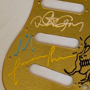 May include: A gold-colored guitar pickguard with multiple signatures in various colors, including white, yellow, and blue. The pickguard has three pickup cutouts and several screw holes.