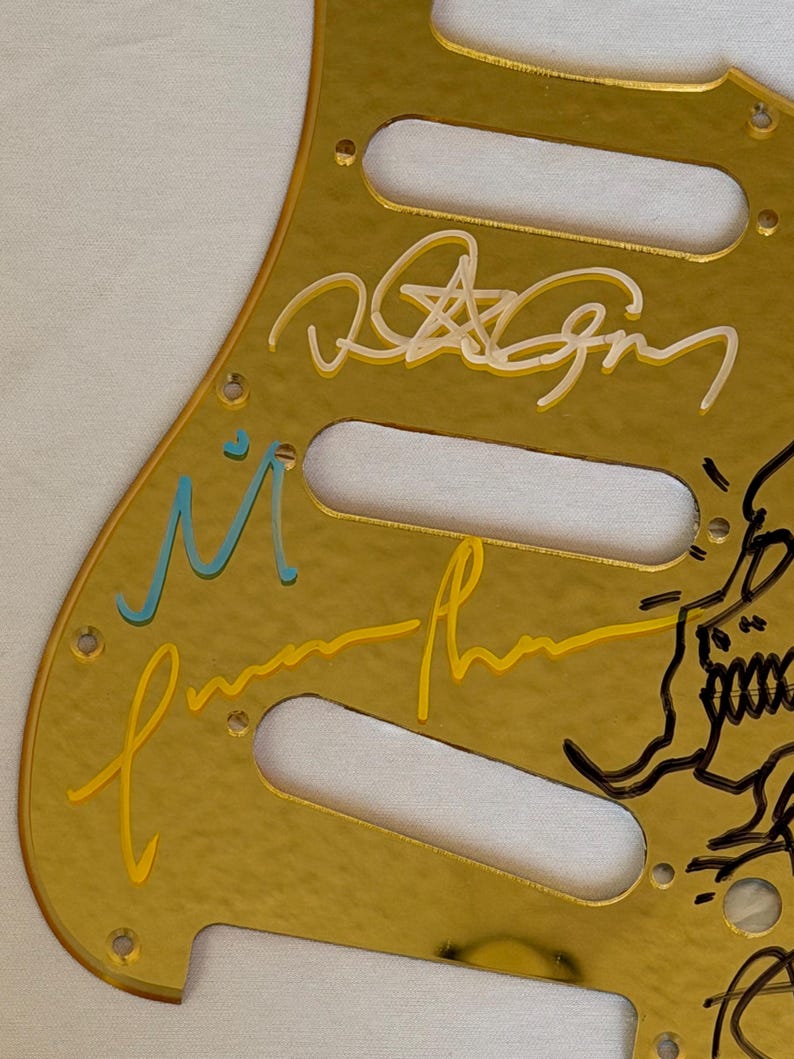 May include: A gold-colored guitar pickguard with multiple signatures in various colors, including white, yellow, and blue. The pickguard has three pickup cutouts and several screw holes.