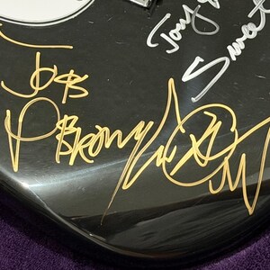 May include: Close-up of a black guitar body with gold and silver signatures. The gold signatures include the names "Joe B" and "P. Brown". The silver signature reads "Tony Sweet". The guitar has a glossy finish.