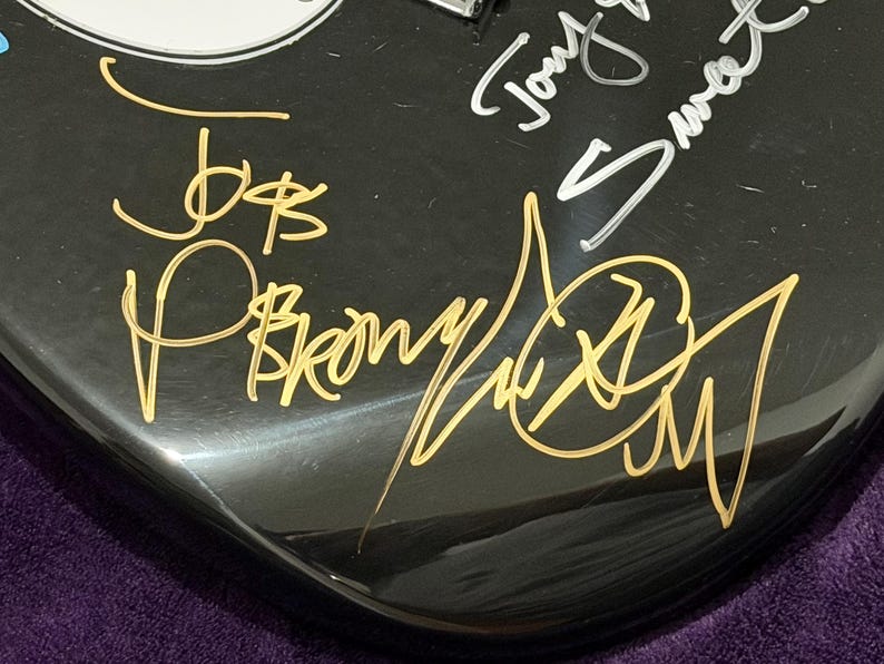 May include: Close-up of a black guitar body with gold and silver signatures. The gold signatures include the names "Joe B" and "P. Brown". The silver signature reads "Tony Sweet". The guitar has a glossy finish.