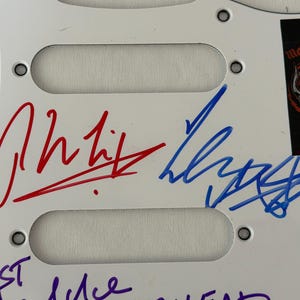 May include: White guitar pickguard with three pickup cutouts. Features signatures in red, blue, and purple ink. A black sticker with the "Mot&ouml;rhead" logo is in the upper right corner. The pickguard has mounting holes.