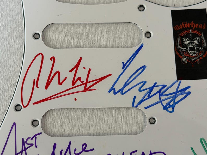 May include: White guitar pickguard with three pickup cutouts. Features signatures in red, blue, and purple ink. A black sticker with the "Mot&ouml;rhead" logo is in the upper right corner. The pickguard has mounting holes.