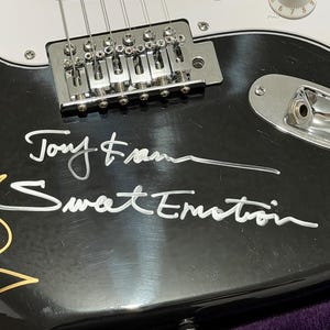 May include: Close-up of a black electric guitar with silver hardware and white accents. The guitar features the signatures "Tony Frank" and "Sweet Emotion" in white script. Gold and white signatures are also visible.
