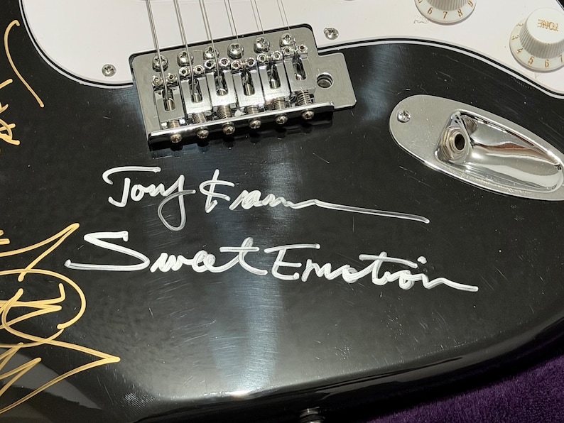 May include: Close-up of a black electric guitar with silver hardware and white accents. The guitar features the signatures "Tony Frank" and "Sweet Emotion" in white script. Gold and white signatures are also visible.