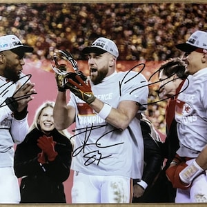 Puede incluir: Un grupo de cuatro personas, incluyendo tres hombres que llevan camisetas grises con las palabras "Kansas City Chiefs" y "Champions" en ellas, están sosteniendo un trofeo. El hombre del centro está sosteniendo el trofeo en alto y sonriendo. El hombre de la izquierda está sosteniendo sus manos juntas frente a él. El hombre de la derecha está sosteniendo un guante rojo y blanco. Una mujer está de pie frente a los hombres, aplaudiendo. El fondo es una multitud de personas.