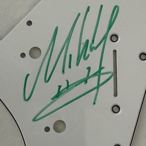May include: White guitar pickguard with green and purple marker signatures. The green signature is the most prominent, with the name "Mikey" visible. The pickguard has several holes and a slot for mounting on a guitar.