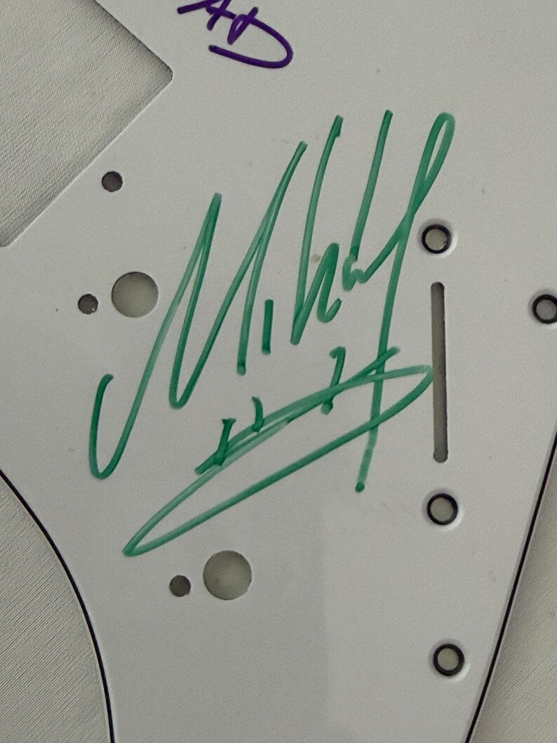 May include: White guitar pickguard with green and purple marker signatures. The green signature is the most prominent, with the name "Mikey" visible. The pickguard has several holes and a slot for mounting on a guitar.