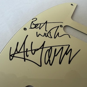 May include: Off-white guitar pickguard with black handwritten text that reads "Best wish" and a signature. The pickguard has several screw holes and a curved shape, likely for a specific guitar model.