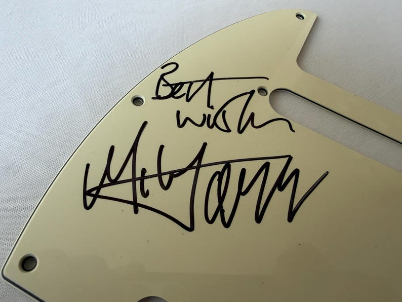 May include: Off-white guitar pickguard with black handwritten text that reads "Best wish" and a signature. The pickguard has several screw holes and a curved shape, likely for a specific guitar model.