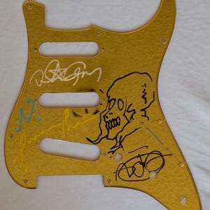 May include: A gold-colored guitar pickguard with a textured surface. The pickguard features multiple signatures in black, white, and yellow ink, along with a black skull illustration. It has three pickup holes and screw holes.