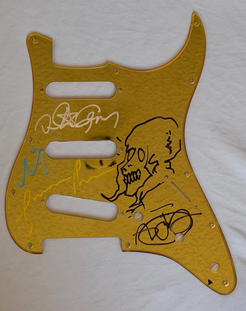 May include: A gold-colored guitar pickguard with a textured surface. The pickguard features multiple signatures in black, white, and yellow ink, along with a black skull illustration. It has three pickup holes and screw holes.