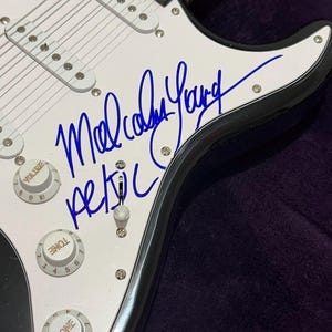 Pu&ograve; includere: Primo piano di una chitarra elettrica bianca con finiture nere. La chitarra presenta una firma in inchiostro blu che recita "Malcolm Young" e il nome della band "AC/DC". La chitarra ha manopole del volume e del tono.