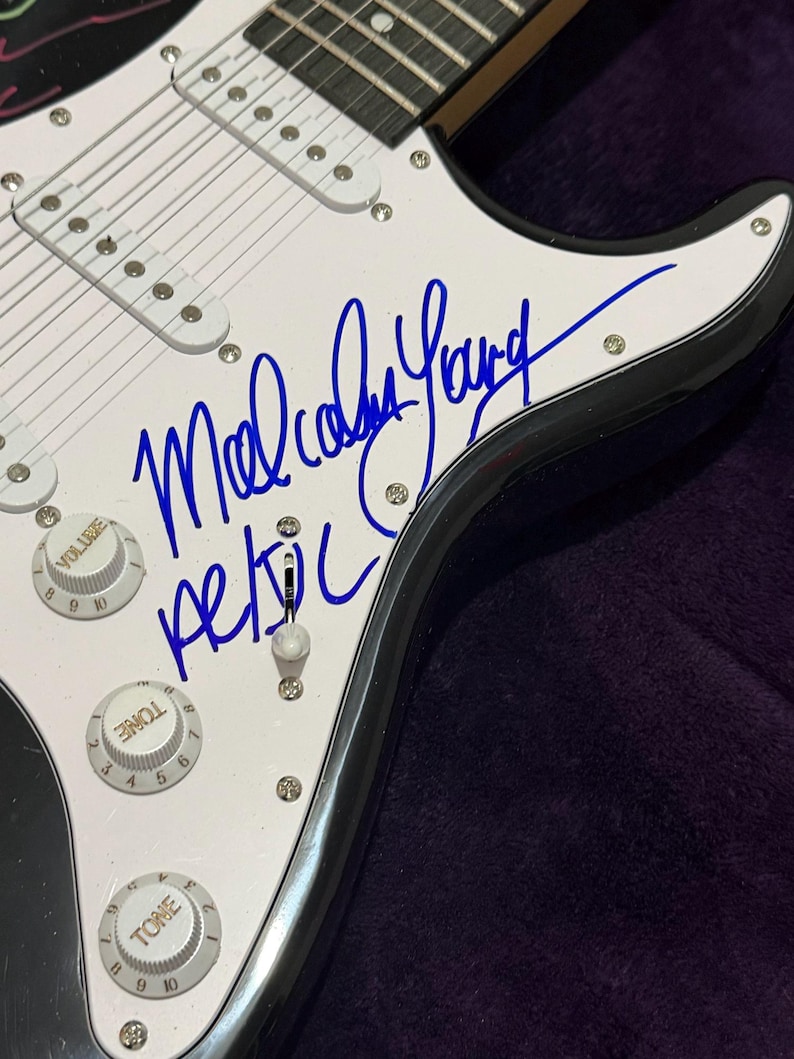 Pu&ograve; includere: Primo piano di una chitarra elettrica bianca con finiture nere. La chitarra presenta una firma in inchiostro blu che recita "Malcolm Young" e il nome della band "AC/DC". La chitarra ha manopole del volume e del tono.