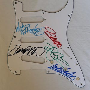 Puede incluir: Golpeador de guitarra blanco con múltiples firmas en tinta azul, roja, verde y negra. El golpeador tiene tres recortes para pastillas y varios agujeros para tornillos. Las firmas parecen ser de una banda o músico.