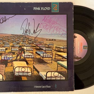 Puede incluir: Portada del álbum "A Momentary Lapse of Reason" de Pink Floyd, con una playa y numerosas camas. La portada incluye firmas y el logotipo de la banda. Se ve un disco de vinilo negro. Álbum de música.