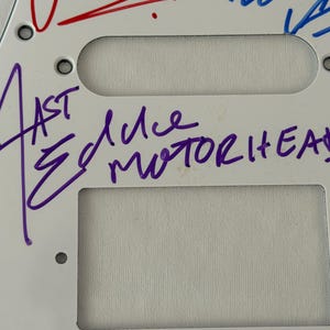 May include: White guitar pickguard with the band name "Motorhead" and signatures in purple and red ink. The pickguard has three rectangular cutouts and several screw holes.