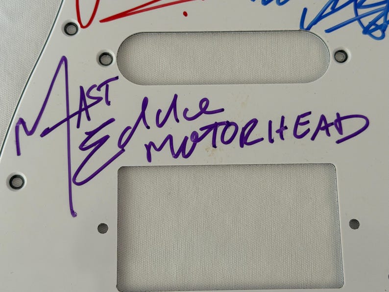 May include: White guitar pickguard with the band name "Motorhead" and signatures in purple and red ink. The pickguard has three rectangular cutouts and several screw holes.