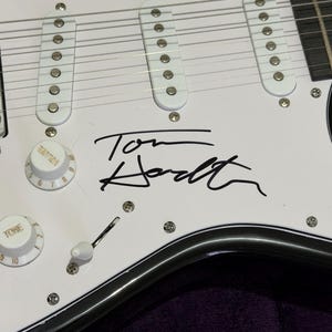 May include: Close-up of a white electric guitar body with black trim. The guitar features white pickups, control knobs, and a signature in black ink that reads "Tom Hodt". The guitar has silver hardware and strings.