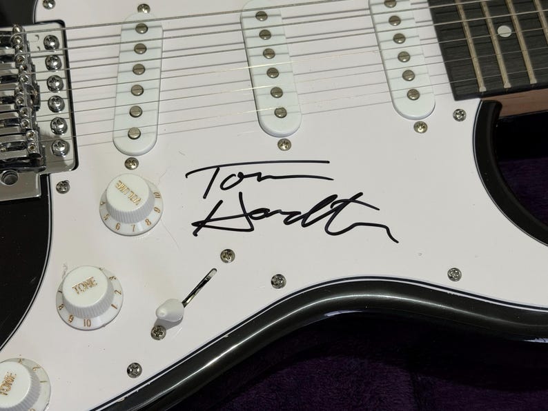 May include: Close-up of a white electric guitar body with black trim. The guitar features white pickups, control knobs, and a signature in black ink that reads "Tom Hodt". The guitar has silver hardware and strings.