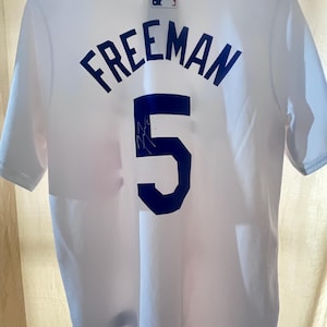 May include: White baseball jersey with the name "FREEMAN" in blue block letters and the number "5" on the back. The jersey has a MLB logo on the upper back. The jersey appears to be made of a breathable fabric.