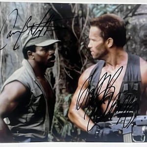 Puede incluir: Fotografía firmada con dos hombres vestidos con ropa militar, con un fondo de jungla. Uno lleva gorra y chaleco, el otro, con un arma, lleva un chaleco táctico. Ambos tienen autógrafos con tinta negra.