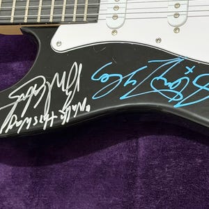 May include: Black electric guitar with white pickguard and silver hardware. The guitar's body is signed in white and blue ink. The signatures are on the lower body of the guitar. The guitar is resting on a purple surface.