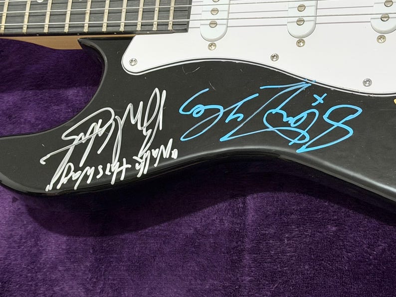 May include: Black electric guitar with white pickguard and silver hardware. The guitar's body is signed in white and blue ink. The signatures are on the lower body of the guitar. The guitar is resting on a purple surface.