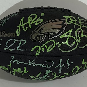 Puede incluir: Balón de fútbol Wilson negro con firmas verdes y plateadas. El logotipo de los Eagles de Filadelfia es visible. El balón tiene el texto "Wilson" y "Philadelphia Eagles".
