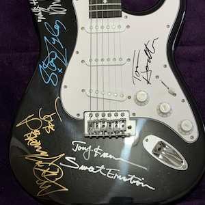 May include: Black electric guitar with white pickguard and silver hardware. The guitar features multiple signatures in various colors, including gold, blue, and white. The guitar has six strings and three white pickups.